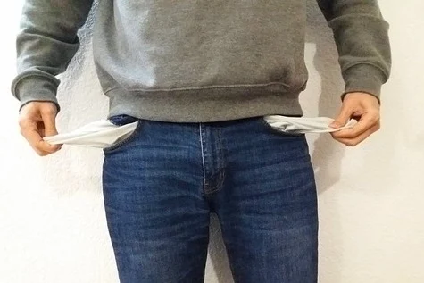 Eine Person steht vor einer hellen Wand und zieht die leeren, nach außen gestülpten Taschen ihrer Jeans hervor.