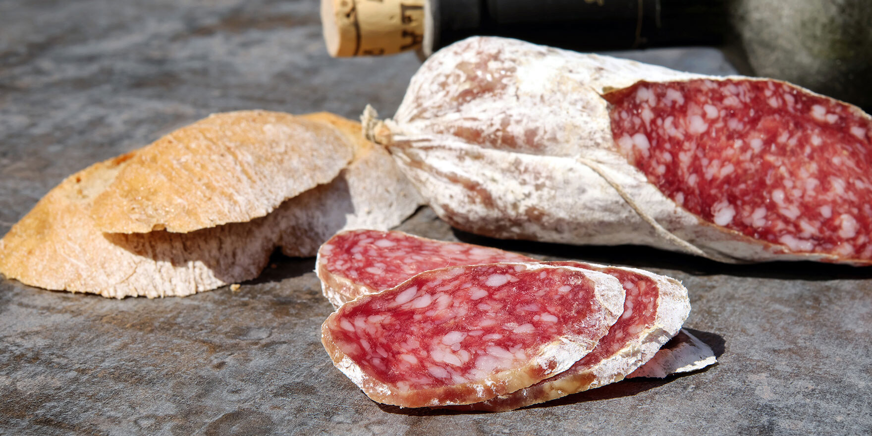 Eine aufgeschnittene Salami liegt neben einem Stück Brot auf dem Tisch, dahinter eine Flasche Wein