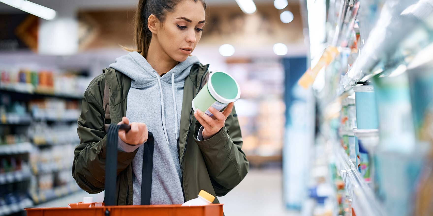 Junge Frau im Supermarkt mit Einkaufskorb, die Milchprodukt in der Hand inspiziert